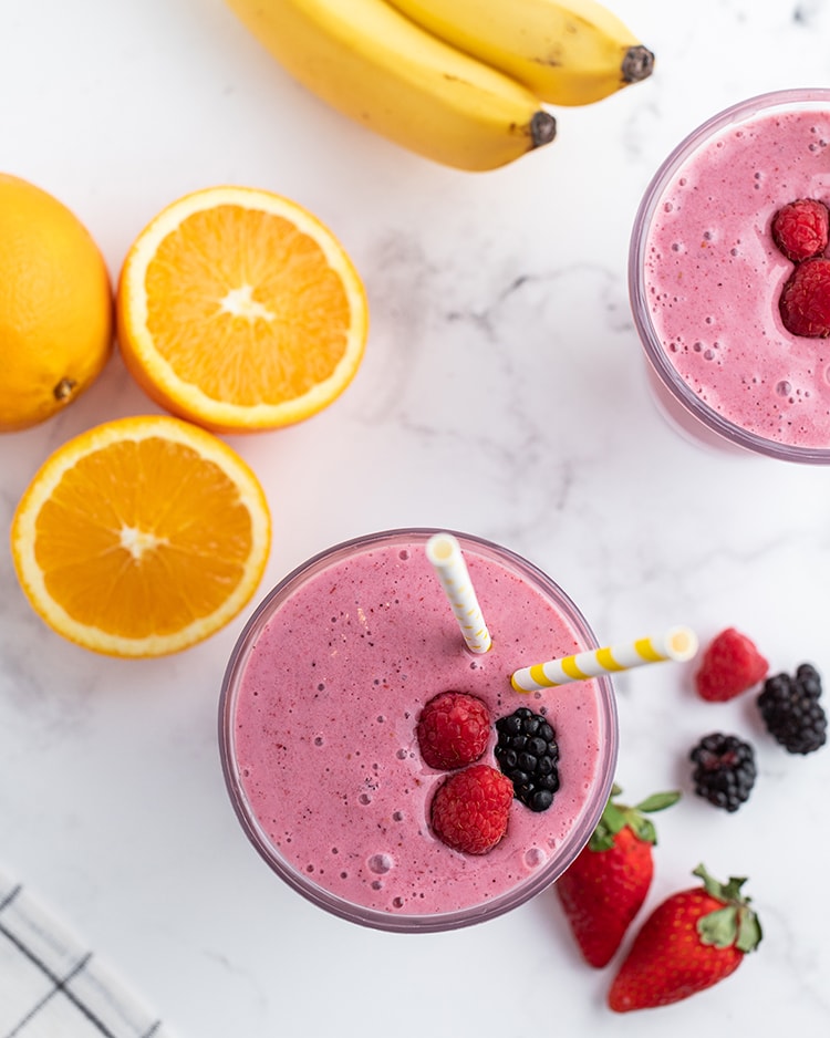 A dark pink smoothie in a glass topped with two raspberries and a blackberry, with two straws, and oranges, bananas, and berries on the side.