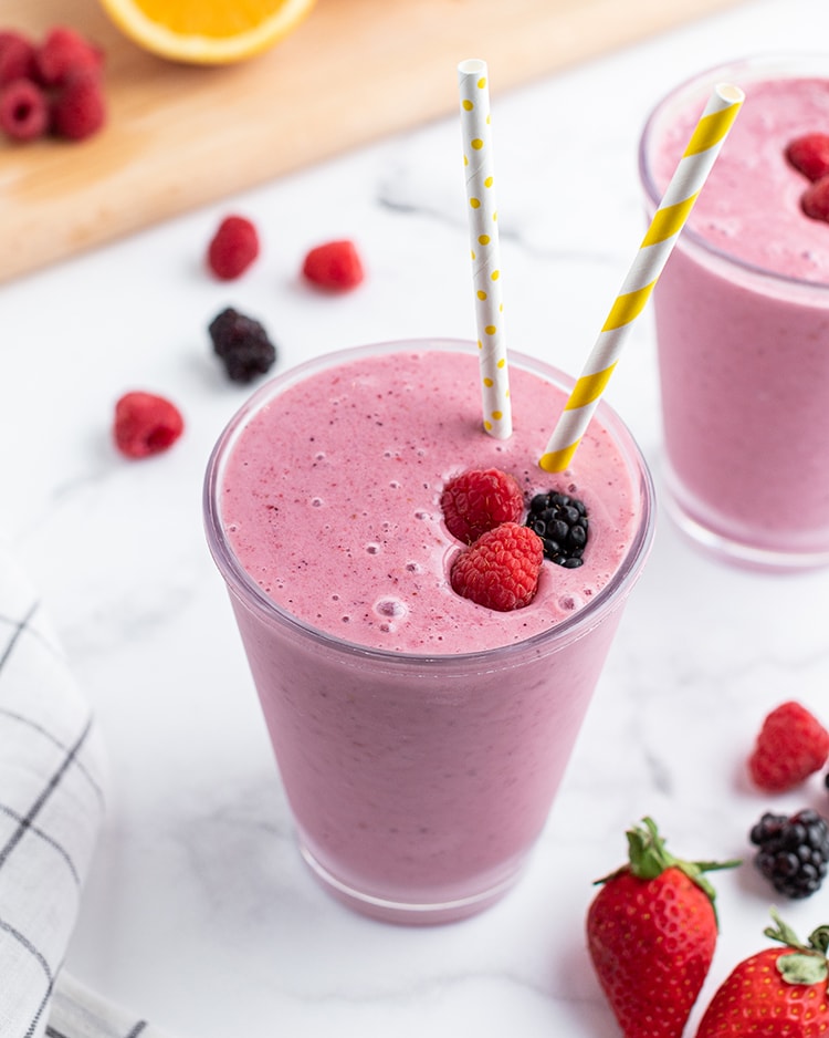 A dark pink smoothie in a glass topped with two raspberries and a blackberry, with two straws, and oranges, bananas, and berries on the side.