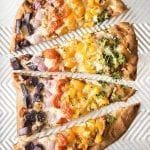 A pizza cut into 4 pieces, and topped with the colors of the rainbow with purple potatoes, red onion, tomatoes, orange and yellow bell pepper and broccoli.
