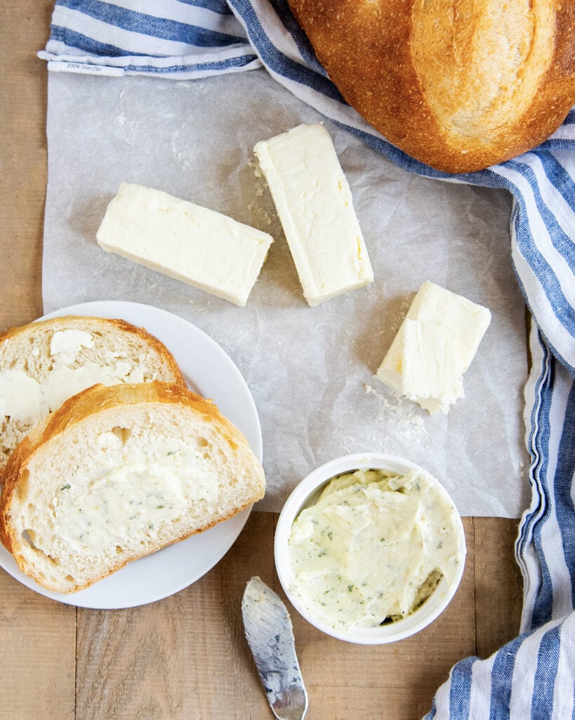 An above photo of sticks of butter on parchment paper, next to a bowl of garlic butter, and butter spread on a piece of bread.