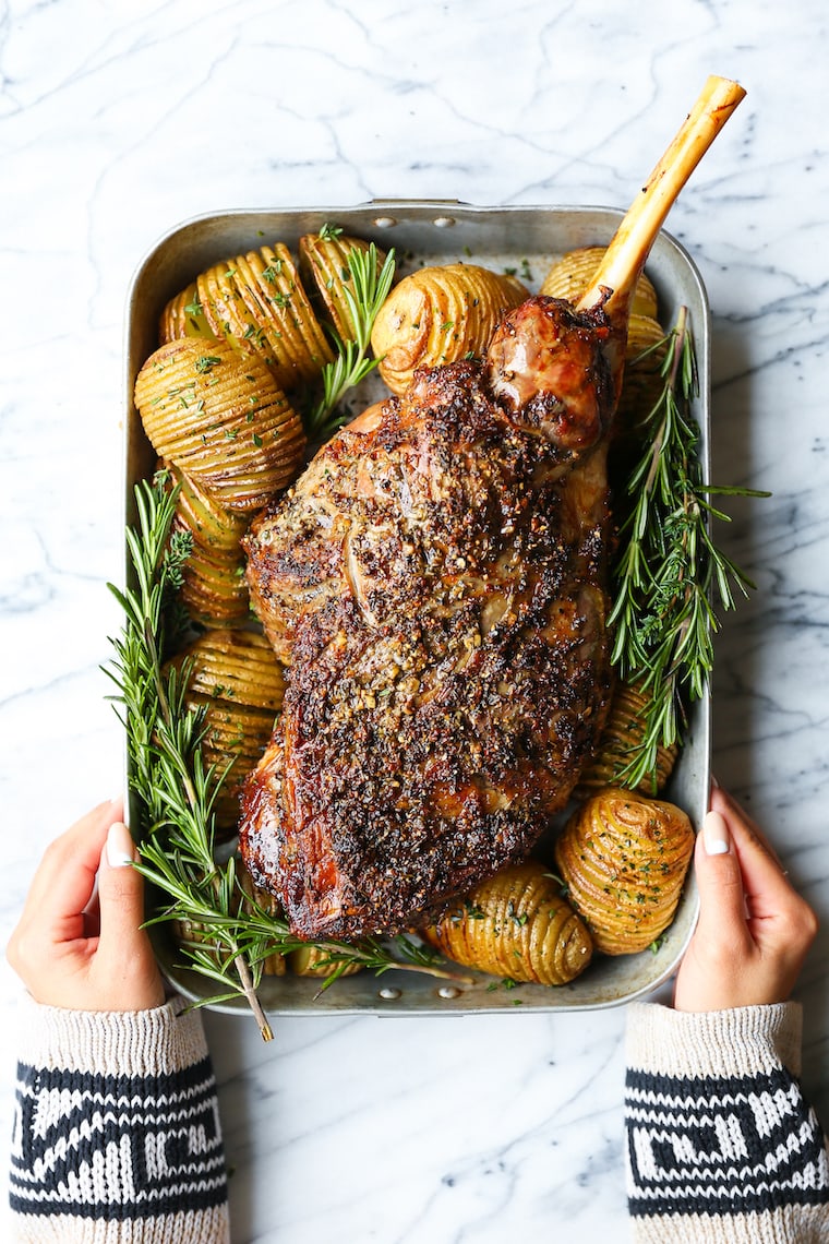 A leg of lamb in a baking pan surrounded by potatoes and rosemary leaves.