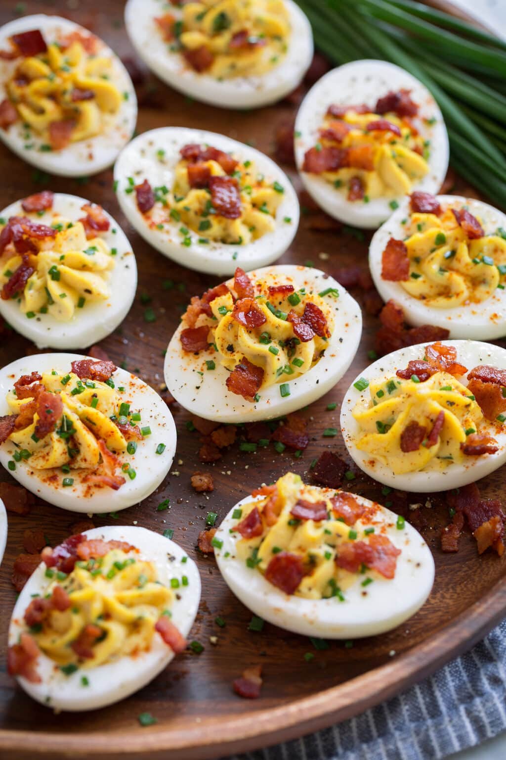 Deviled eggs on a wooden plate, topped with bacon bits and chopped up chives and sprinkled with paprika.