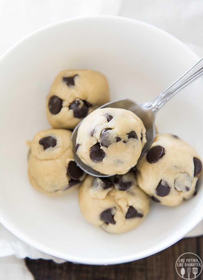 A bowl of 6 small balls of cookie dough. There are lots of chocolate chips peaking through the cookie dough. One of the balls is on a spoon.