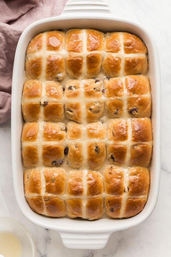 Rows of hot cross buns in a baking pan, you can see the white cross on them and some raisins peaking through.