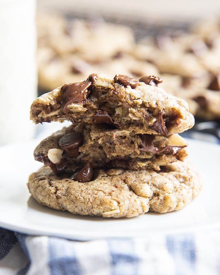 A stack of chocolate chip cookies, the top cookie is broken in half to show the melted chocolate chips and oats in the middle.