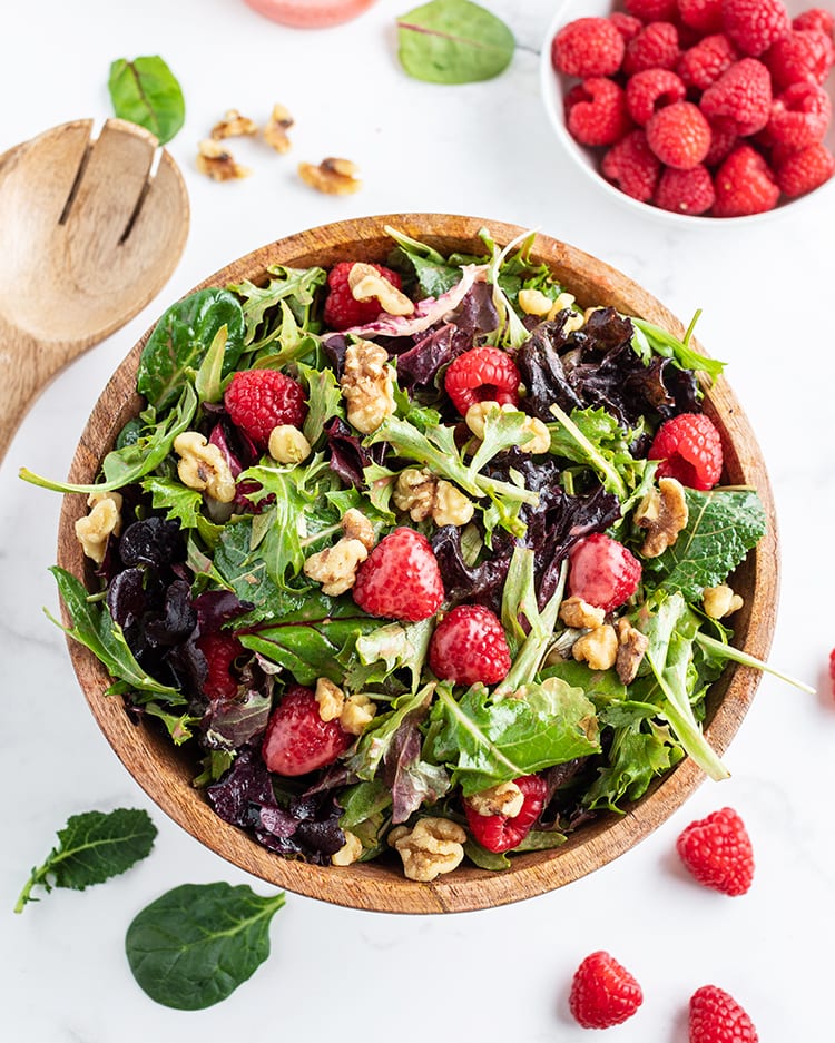 A salad in a wooden bowl with spring mix lettuce, raspberries, and walnuts, with a faint dressing over the salad.