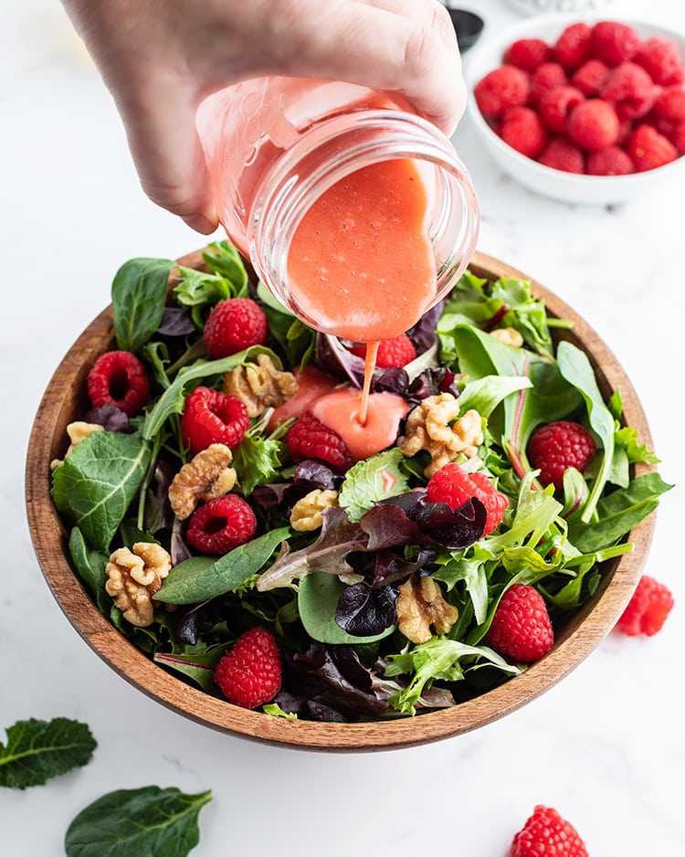 Raspberry vinaigrette being poured out of jar onto a salad with raspberries, walnuts, and spring mix lettuce.