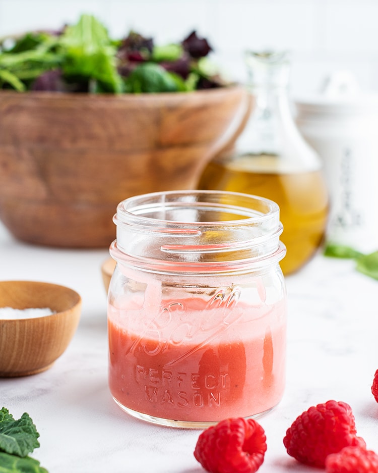 A jar half full of homemade raspberry vinaigrette. There is a bowl of salad behind it.