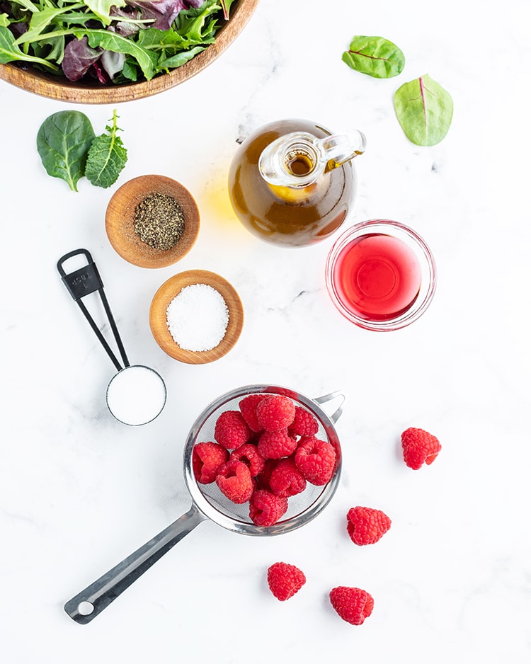 The ingredients needed to make homemade raspberry vinaigrette. A bottle of olive oil, a small bowl of red wine vinegar, raspberries, sugar, salt, and pepper. 