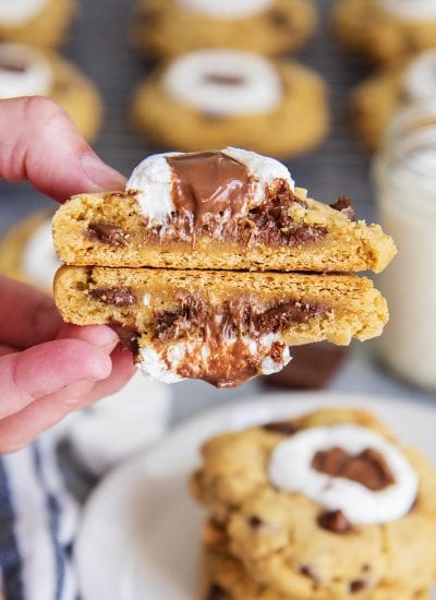 A hand holding two halves of a s'mores cookie showing the gooey inside of the cookie, and graham cracker on the bottom.