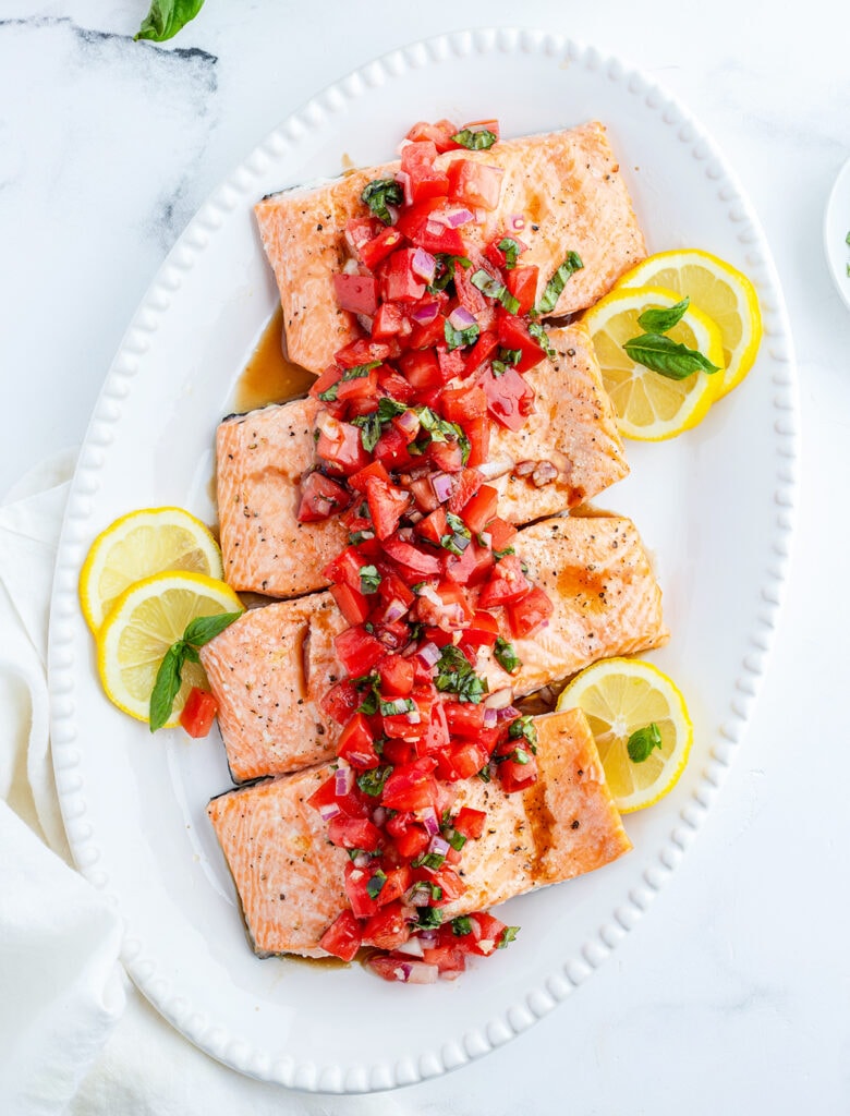 A top view of four pieces of salmon with tomato basil relish, and lemon slices.