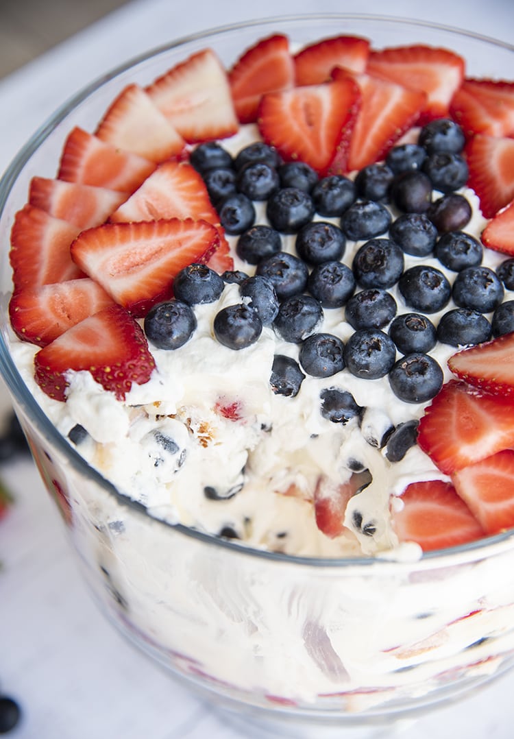Red white and blue trifle displayed in a glass display bowl with a scoop taken out and berries on top.