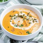 A bowl of tomato and squash soup topped with cream, pumpkin seeds, and fresh parsley,