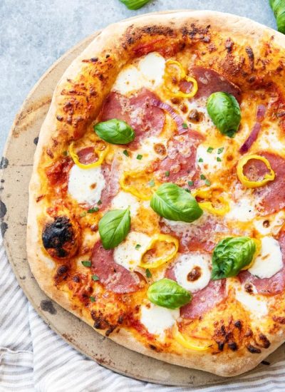 An overhead photo of a salami pizza topped with banana peppers, red onions, mozzarella pears, and fresh basil.