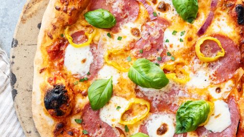 An overhead photo of a salami pizza topped with banana peppers, red onions, mozzarella pears, and fresh basil.