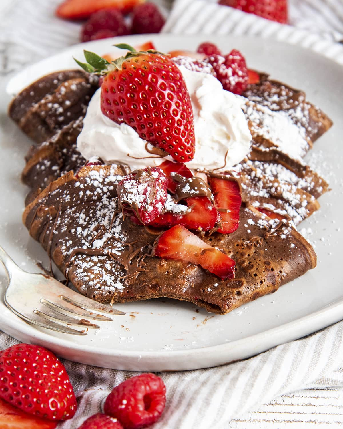 A plate of folded up chocolate crepes with a bite missing from one in the front.