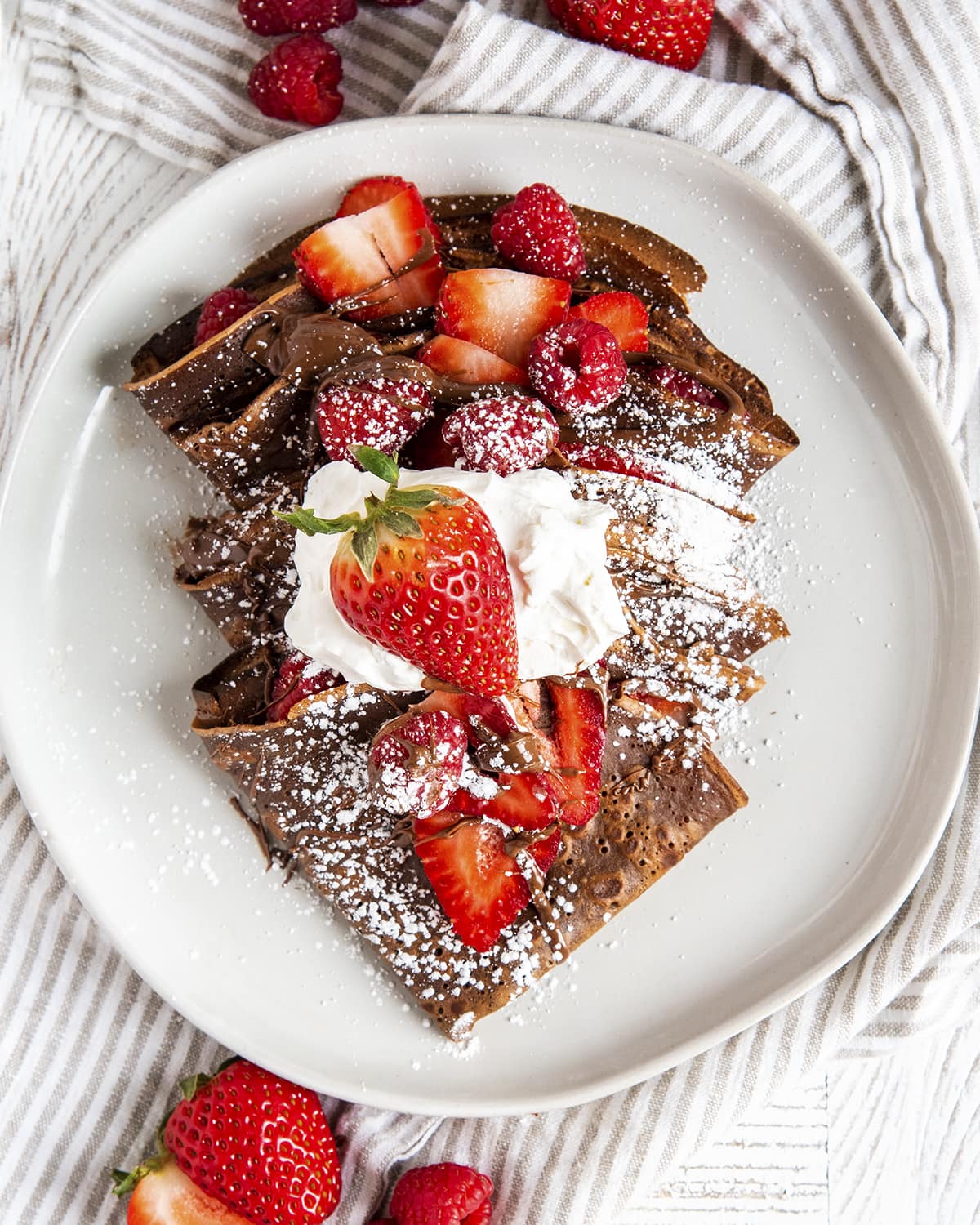 An overhead photo of chocolate crepes with strawberries, powdered sugar, and whipped cream on a plate.