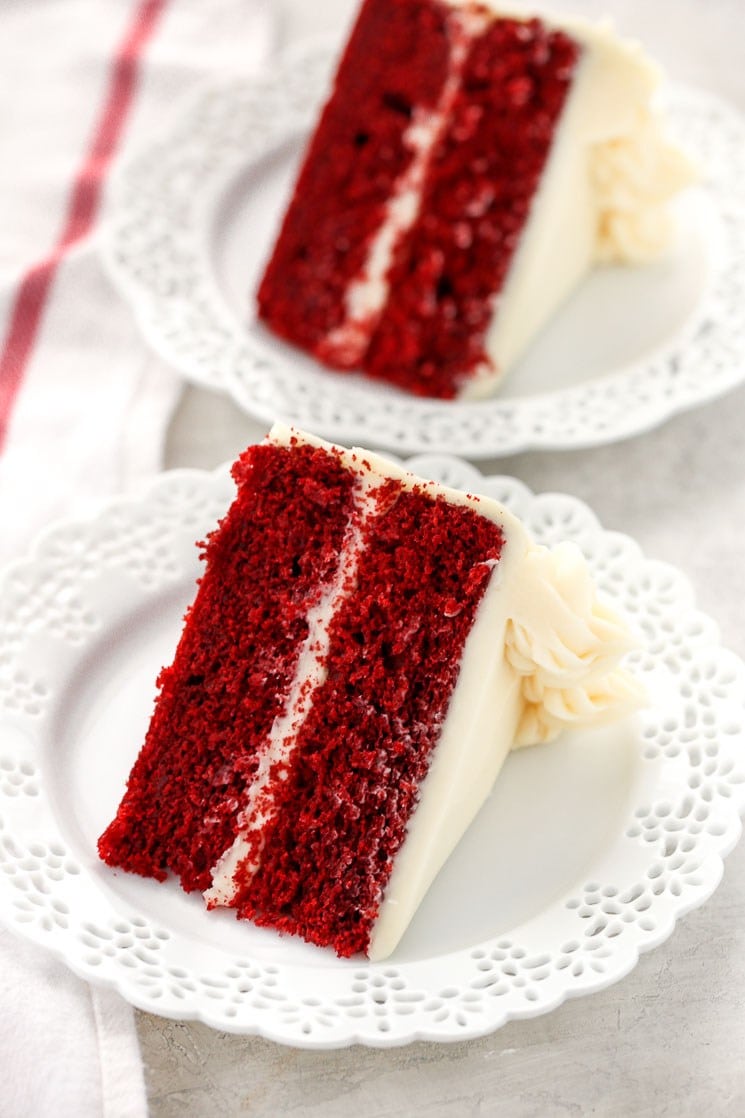 A slice of red velvet cake with cream cheese frosting on a plate, with another slice behind