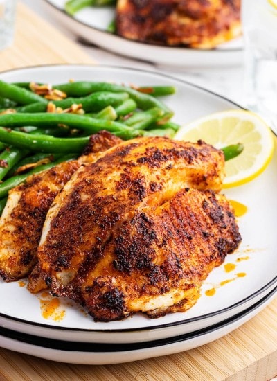 Two pieces of blackened tilapia on a plate with green beans behind it.