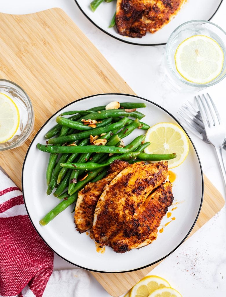 An overhead shot of blackened tilapia on a plate with green beans, and a lemon slice.