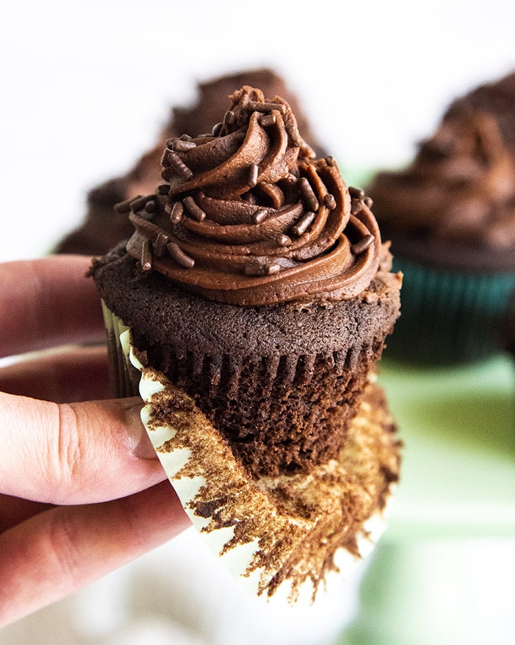 A hand holding a partially unwrapped chocolate cupcake with chocolate frosting. 