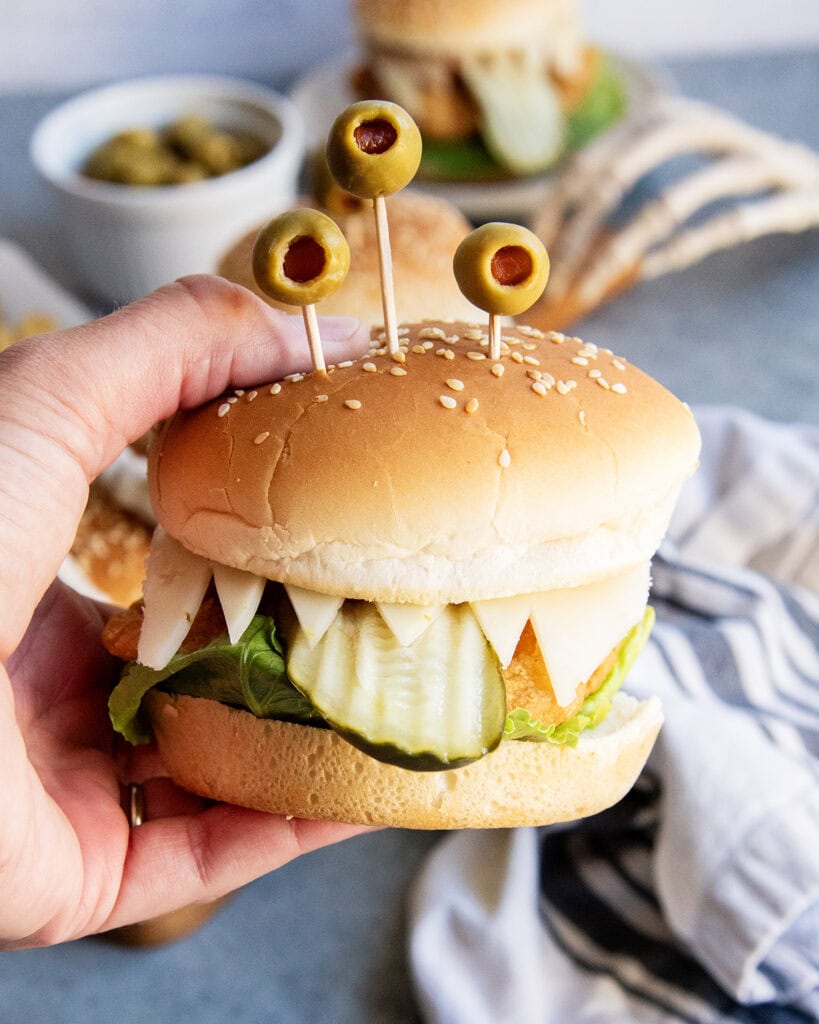 A hand holding a chicken sandwich monster topped with olive "eyes" and the cheese is cut to look like teeth.
