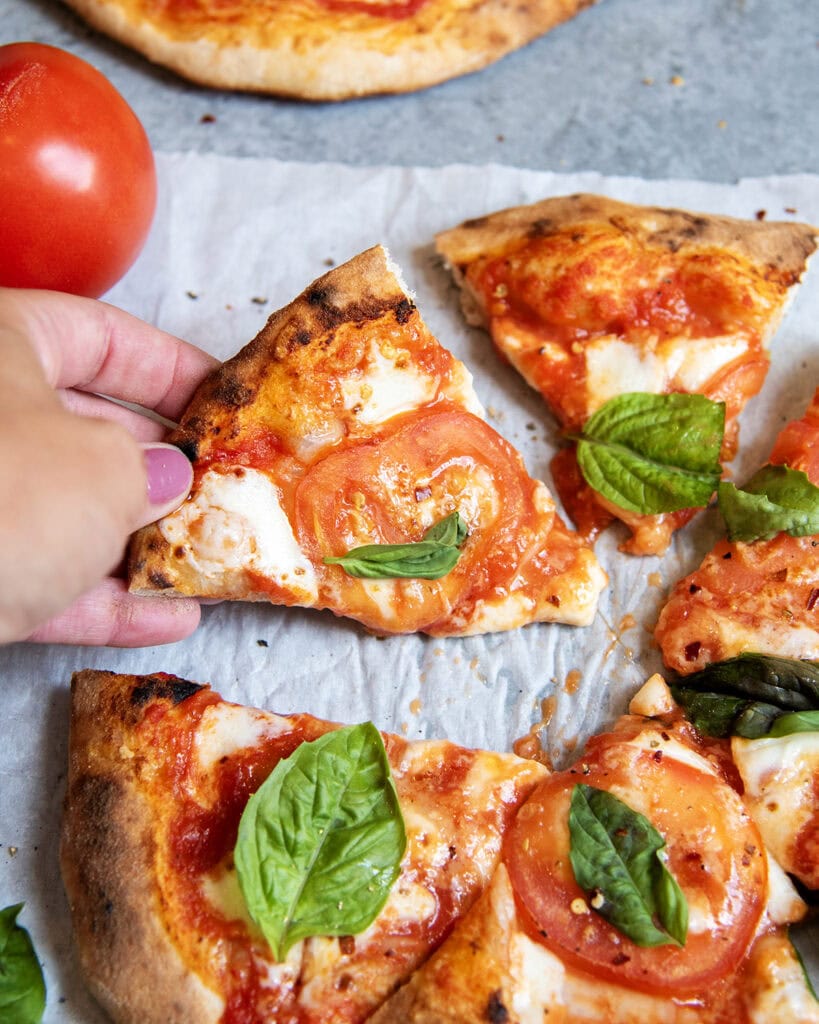 A hand grabbing a slice of Margherita Pizza topped with fresh basil.