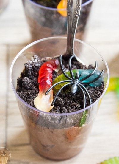 A cup of pudding topped with crushed Oreo cookies and gummy worms, and a spoon in the cup.