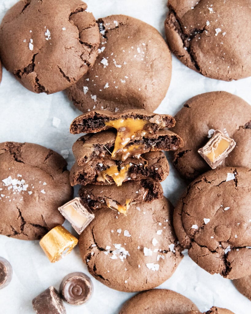 An above view of four halves of chocolate cookies, with a gooey rolo caramel center, next to a pile of more chocolate cookies.