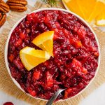 A bowl of cranberry sauce topped with a couple small orange slices with a spoon in the bowl.