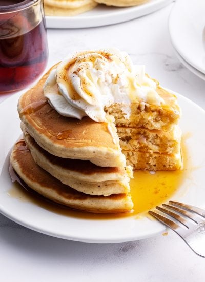 A stack of eggnog pancakes on a plate topped with whipped cream, and syrup with a cut taken out of it showing the middle of the pancakes.