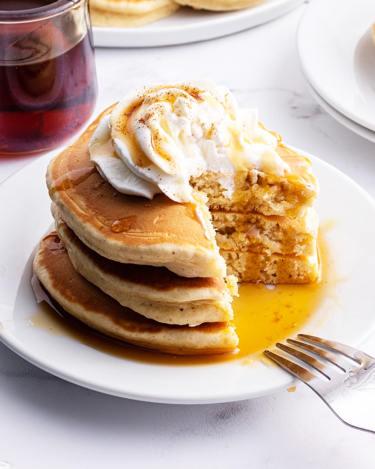 A stack of eggnog pancakes on a plate topped with whipped cream, and syrup with a cut taken out of it showing the middle of the pancakes.
