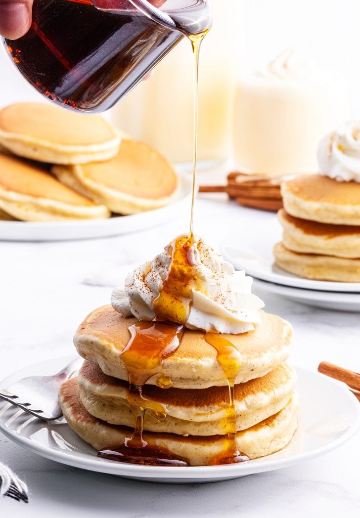 Syrup being poured over a stack of pancakes topped with whipped cream.