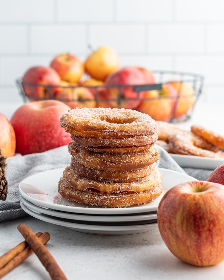 A stack of apple rings coated in cinnamon sugar and set on a plate with a basket of apples in the back