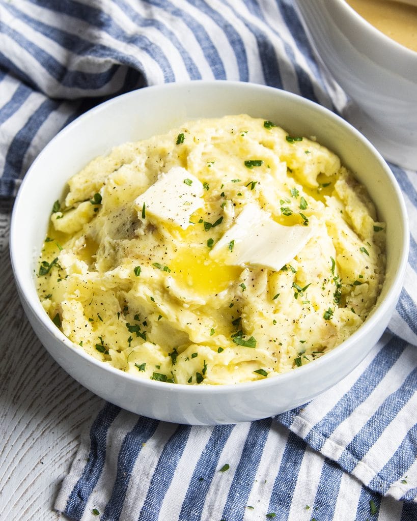 A bowl of golden mashed potatoes topped with butter, on top of a blue and white striped cloth.