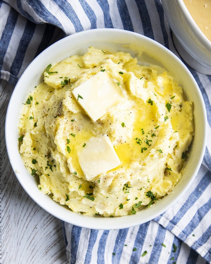 An overhead photo of a bowl of golden mashed potatoes topped with butter, and fresh parsley.