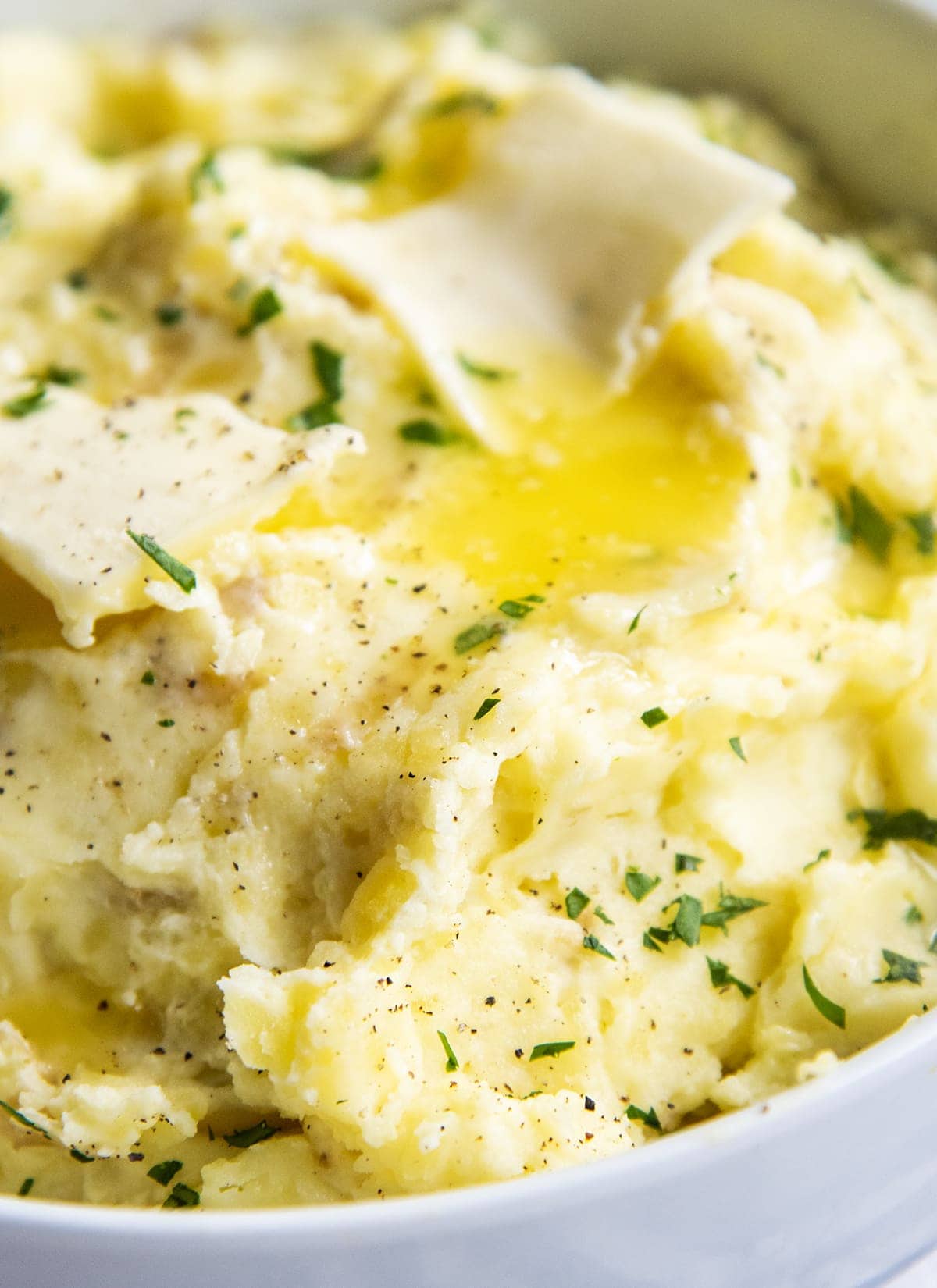 A close up of mashed potatoes in a white bowl topped with melted butter, butter pats, black pepper, and fresh parsle.y