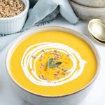 A bowl of butternut squash soup topped with a ting of cream, sunflower seeds, and two small sage leaves.