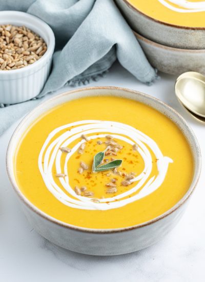 A bowl of butternut squash soup topped with a ting of cream, sunflower seeds, and two small sage leaves.