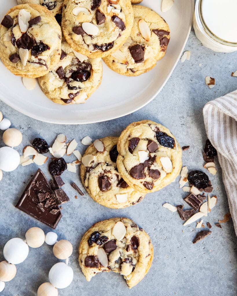 Cherry almond chocolate chip cookies on a plate, and next to the plate on a counter top, one cookie has a bite out of it.