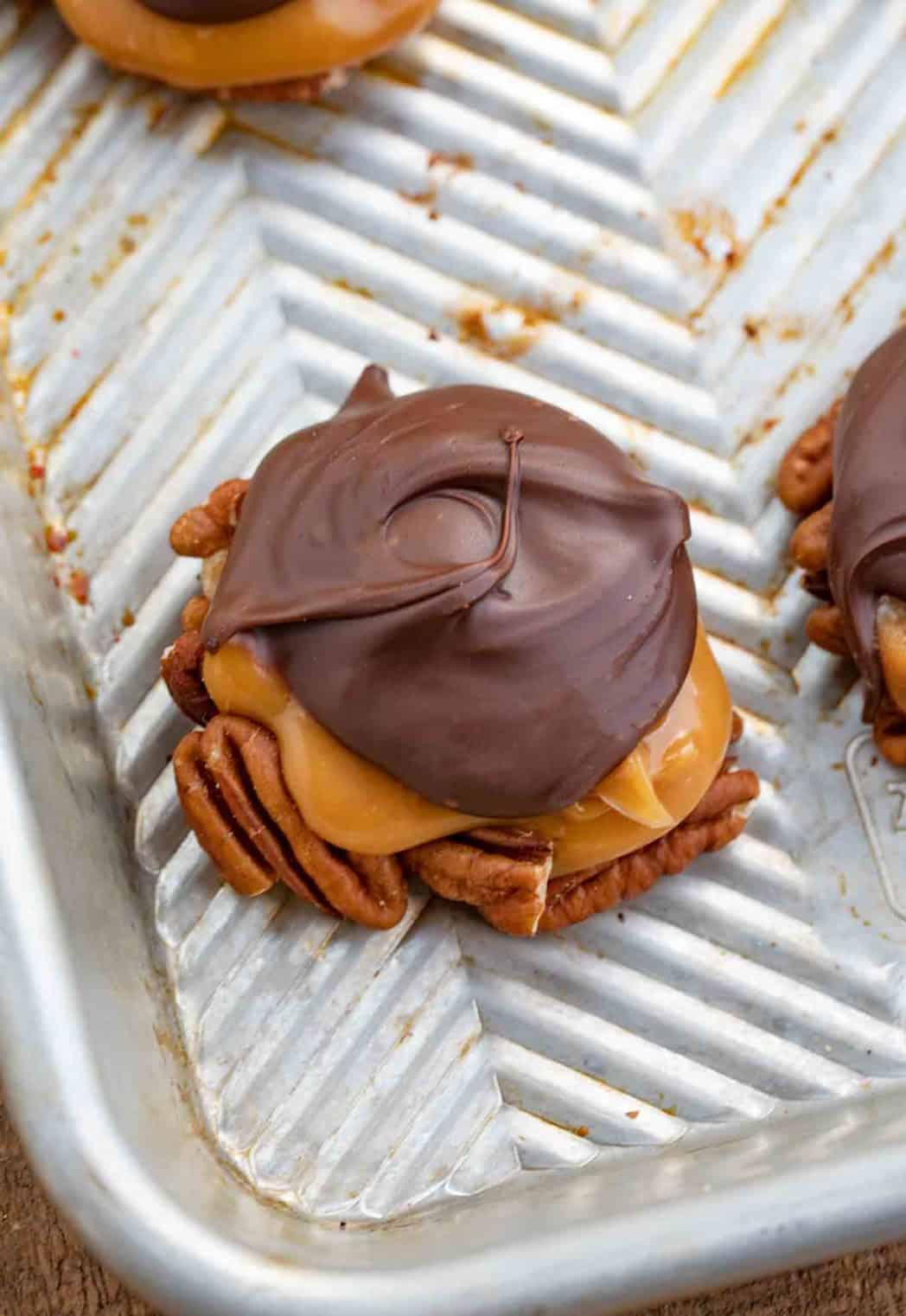 A homemade turtle on a cookie sheet, showing pecans on the bottom, caramel in the middle, and chocolate swirled around on top.