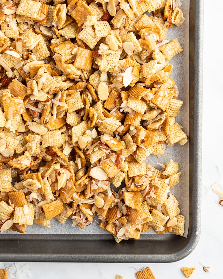 Gooey coconut chex mix on a cookie sheet. It has rice chex, golden grahams, almonds, and coconut.