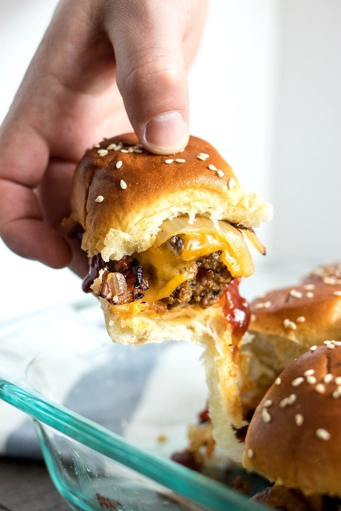 A slider being lifted out of a pan. It has meat, bbq sauce, onions, and cheddar cheese on it.