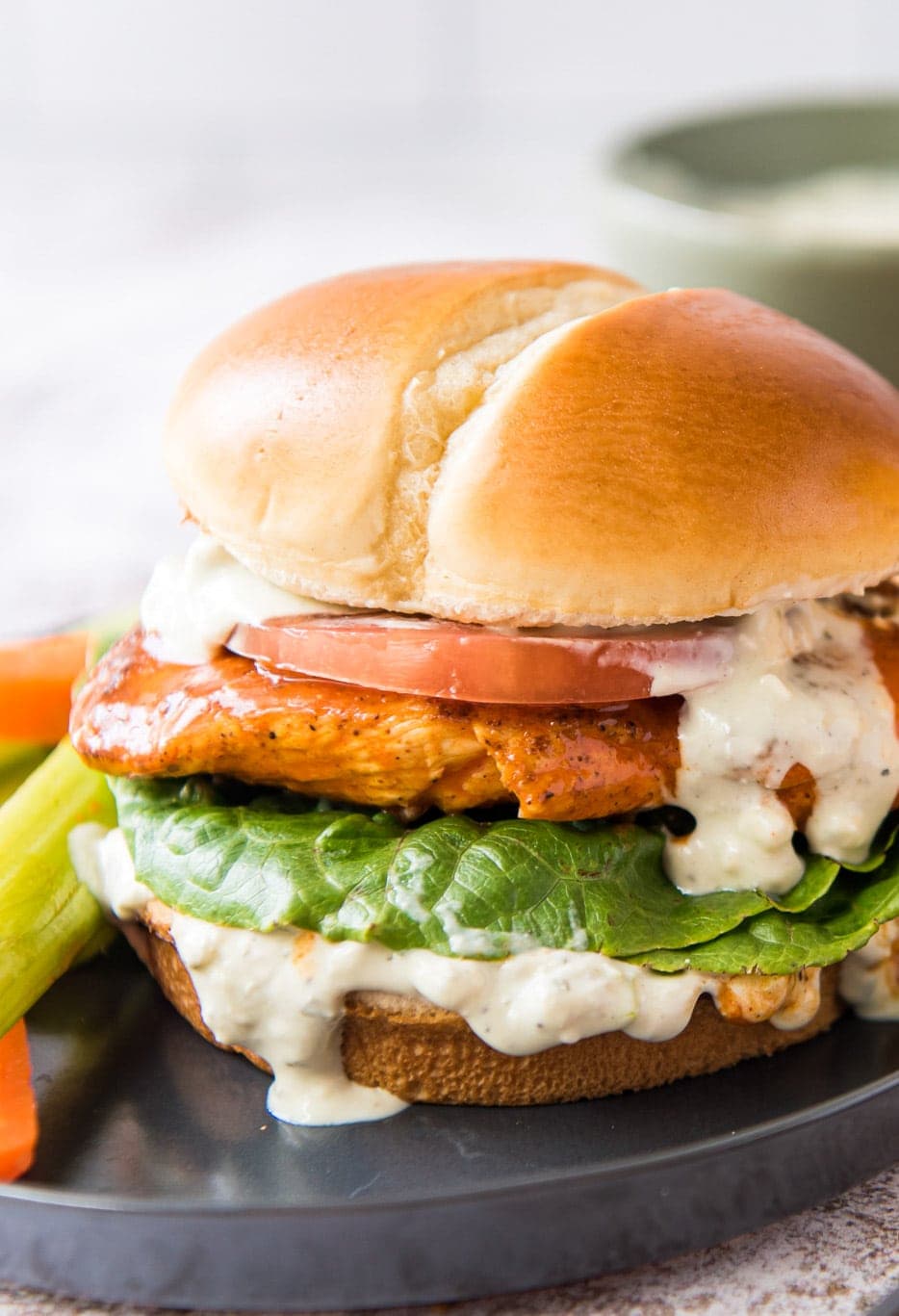 A chicken sandwich with a creamy white sauce, lettuce, a chicken breast with buffalo sauce, and a slice of tomato, on a bun.