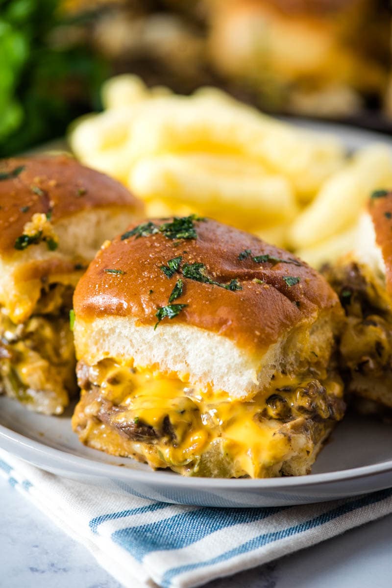 A cheese steak slider on a plate, you can see some flank steak covered in melty cheese and the bun is topped with butter and parsley.