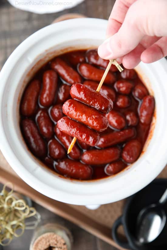 Three little smokies on a toothpick being held by a hand above a slow cooker full of little smokies.
