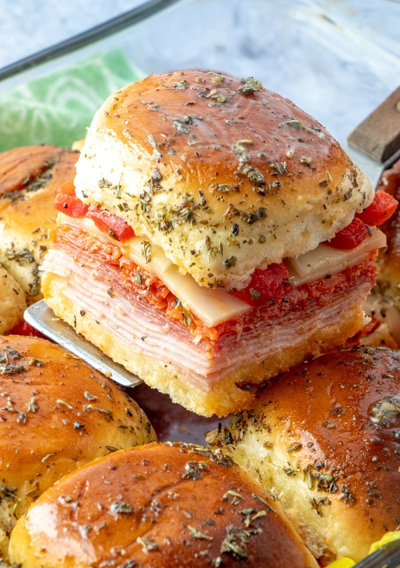 A slider sandwich full of ham, salami, tomatoes, and cheese, all topped with herby butter.