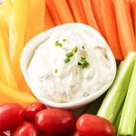 A close up shot of ranch dip in a small white bowl with vegetables surrounding it.