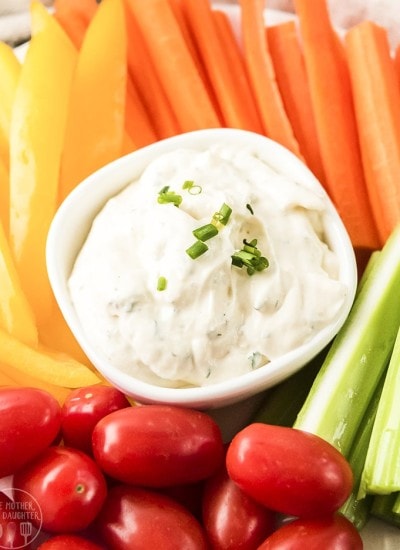 A close up shot of ranch dip in a small white bowl with vegetables surrounding it.