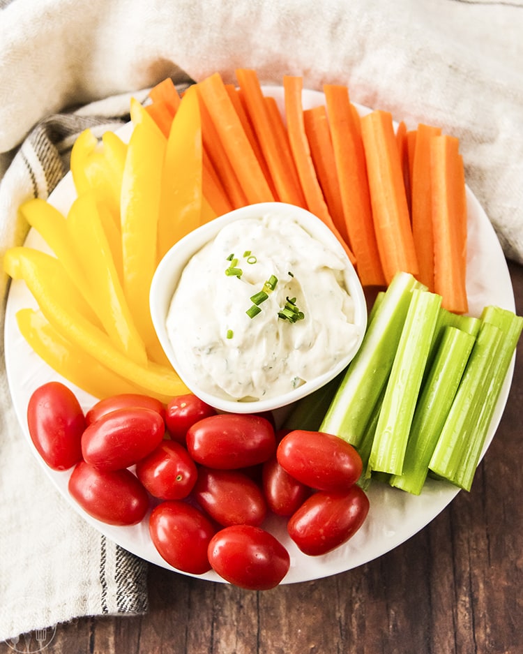 A close up shot of ranch dip in a small white bowl with vegetables surrounding it.